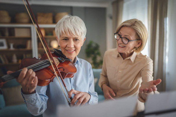Two women mature female teacher give private violin lesson to her student or friend at home help her to learn to play in room hobby and leisure active senior concept copy space - Foto, immagini
