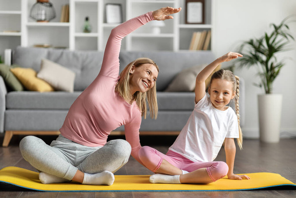 Ευτυχισμένη νεαρή μητέρα και η μικρή κόρη της που κάνουν σπορ μαζί στο σπίτι, χαρούμενη μαμά και χαριτωμένο θηλυκό παιδί κάνοντας side Bend ασκήσεις, εκπαίδευση στο στρώμα γυμναστικής στο εσωτερικό του σαλονιού - Φωτογραφία, εικόνα
