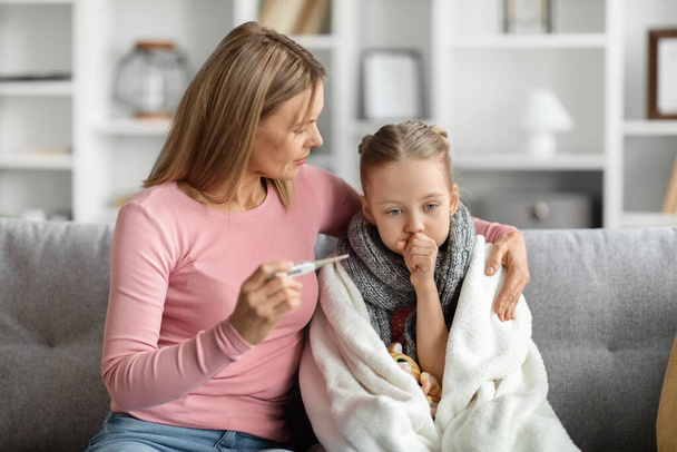 Flu Concept. Worried mother checking her daughters temperature at home, caring mom holding thermometer and embracing sick little girl covered in blanket, female kid coughing into fist - Foto, immagini