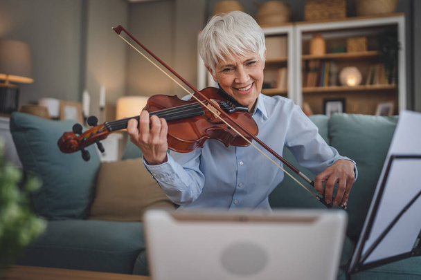 Mature senior caucasian woman learn to play violin practice at home online professor teacher teach or have internet private class on digital tablet while sit on sofa bed at home active senior hobby - Photo, Image