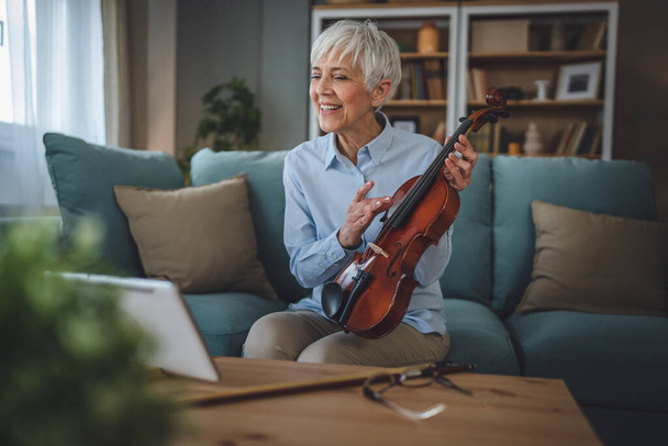 Mature senior caucasian woman learn to play violin practice at home online professor teacher teach or have internet private class on digital tablet while sit on sofa bed at home active senior hobby - Fotografie, Obrázek