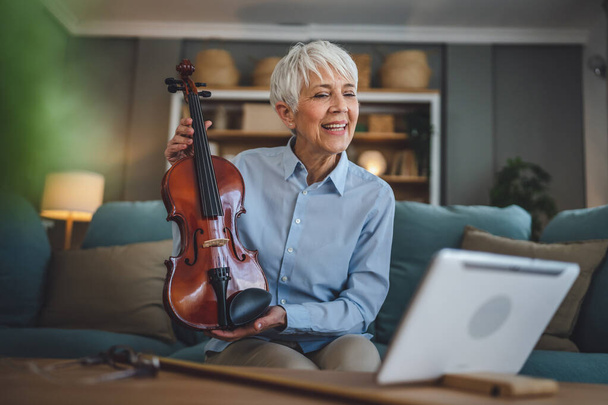 Ώριμη ηλικιωμένη καυκάσια γυναίκα μαθαίνουν να παίζουν βιολί πρακτική στο σπίτι σε απευθείας σύνδεση καθηγητής δάσκαλος διδάσκουν ή να έχουν ιδιωτικό internet τάξη σε ψηφιακή ταμπλέτα, ενώ κάθονται σε καναπέ-κρεβάτι στο σπίτι ενεργό ανώτερος χόμπι - Φωτογραφία, εικόνα