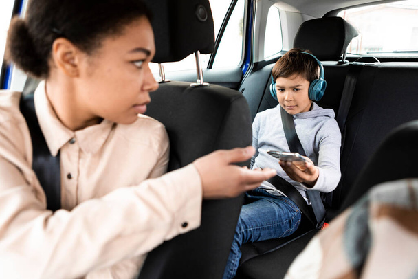 Angry black woman taking back smartphone from little son in car. Family, transport, road trip, travel and people concept - Photo, Image