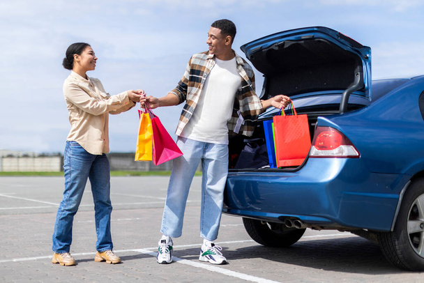 Junges afrikanisch-amerikanisches Paar legt Einkaufstüten aus dem Auto, nachdem es in einem Einkaufszentrum eingekauft hat, Mann und Frau schauen einander an und lächeln in voller Länge - Foto, Bild