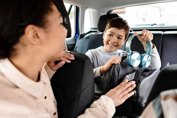 Happy european boy putting headphones in his backpack and smiling to mother while sitting on backseat inside electric car, mother looking at son - Photo, Image