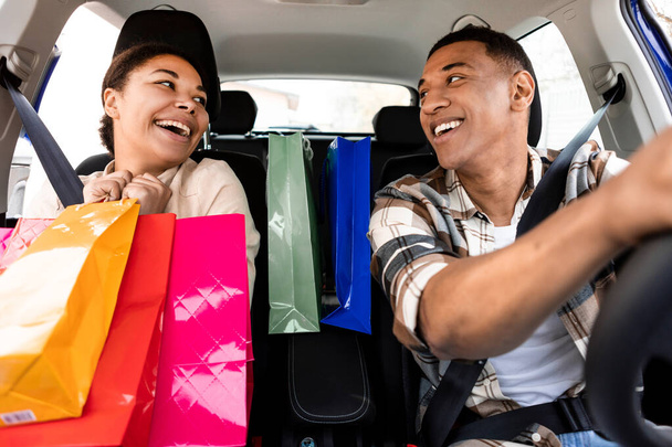 Izgalmas fekete házastársak ülnek az autóban élvezi lovagolni sikeres vásárlás után, nő kezében színes vásárlói táskák, egymásra néz, és mosolyog - Fotó, kép