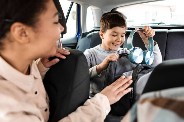 Happy boy packing his backpack and putting headphones inside while sitting on backseat inside electric car, mother looking at son and smiling - Photo, Image