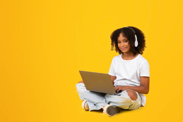 Happy teen curly black girl, headphones typing on laptop, watch video lesson, play online game, sit on floor, isolated on yellow studio background. Homework, study, knowledge with device - Photo, Image