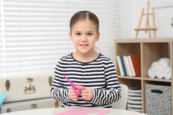 Cute little girl with scissors and pink paper at desk in room. Home workplace - Foto, Imagen