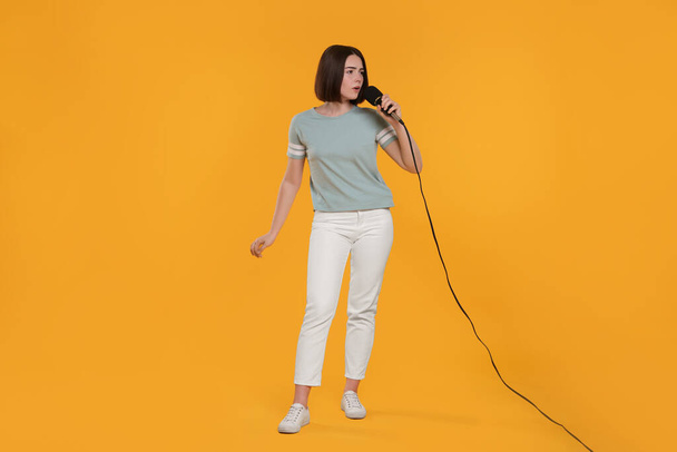 Beautiful young woman with microphone singing on yellow background - Foto, afbeelding