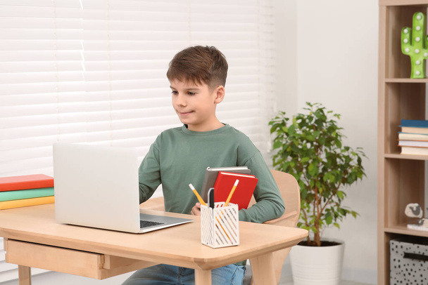 Boy with books using laptop at desk in room. Home workplace - 写真・画像
