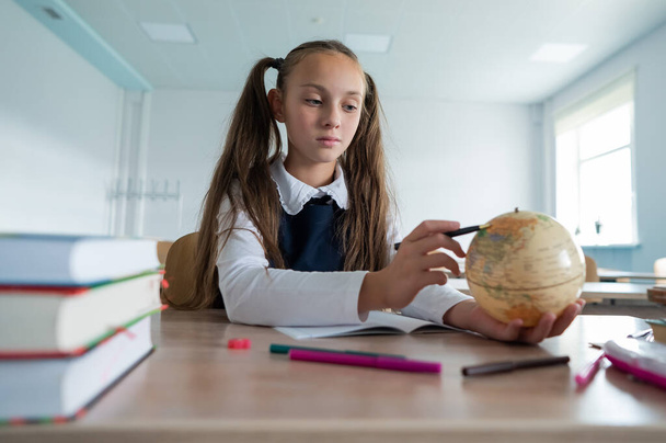 Caucasian schoolgirl sits at her desk at school and studies the globe - Foto, Imagen