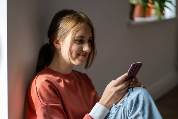 Happy smiling gen Z teen girl relax sitting on floor receiving message on smartphone reading text in screen surfing social media. Interested teenager using online virtual app play game on mobile phone - Foto, Imagen