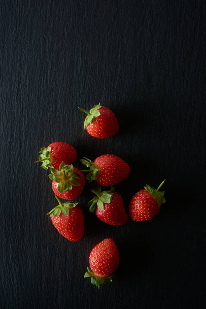 strawberries on black textured background, bright red color popular juicy fruit with sweet taste and fragrant aroma, healthy and highly nutritious culinary ingredient, taken from above with copy space - Photo, image
