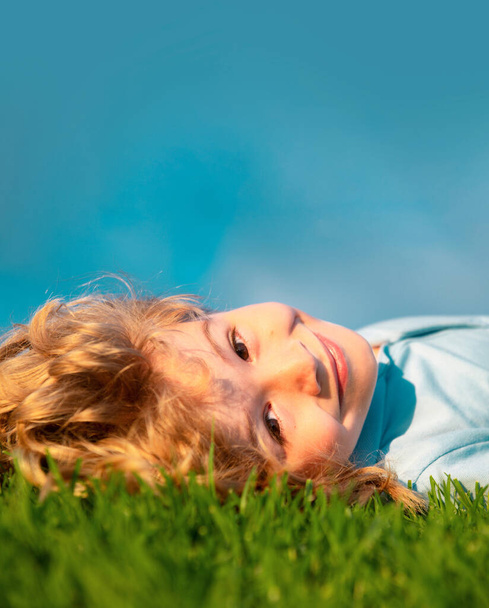 Gelukkig kind genietend op grasveld en dromend. Kind ontspannen op de natuur. Kopieer ruimte op blauwe lucht. Close up portret van grappige kinderen gezicht - Foto, afbeelding