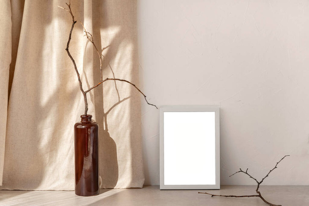 Empty picture frame mockup standing on a stone tabletop, brown vase with wooden branch, sunlight shadows on linen curtain and neutral beige wall background, minimalist design template, copy space - Photo, Image