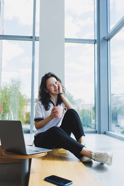 Uśmiechnięta kobieta z kręconymi włosami siedząca w loftowej kawiarni, pracująca przy komputerze, czytająca wiadomości na smartfonie. Praca zdalna - Zdjęcie, obraz