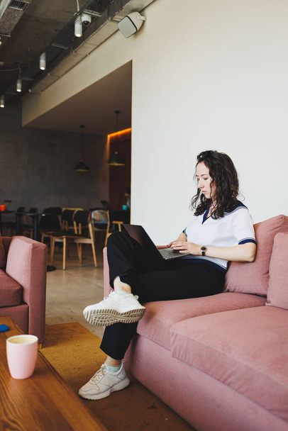 ラップトップや電話がオンラインで働いているテーブルで、モダンなカフェの快適なソファに座ってカジュアルな服のきれいな女性。リモートワーク - 写真・画像