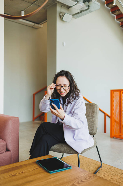 Emocjonalne ładna kobieta w okularach w ubraniach casual siedzi w wygodnym fotelu w nowoczesnej kawiarni przy stole z telefonem pracy online. Praca zdalna - Zdjęcie, obraz