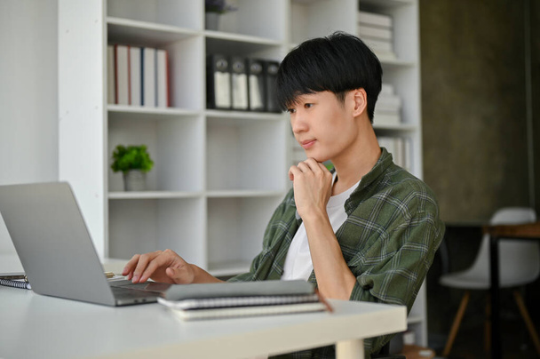 A smart and professional young Asian male office worker or assistant sits at his desk in a modern office and focuses on his work on his laptop computer. - Fotó, kép