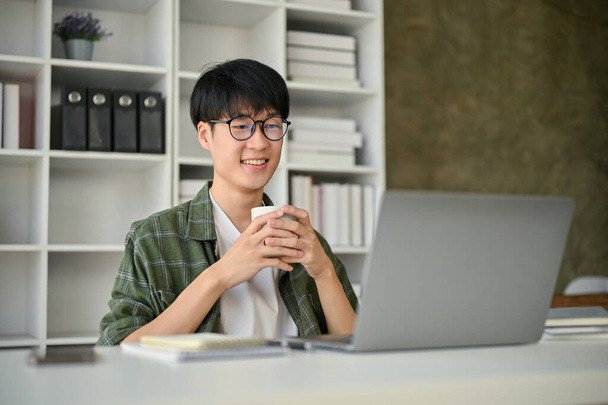 A smart and happy young Asian male office worker or assistant in glasses sits at his desk and holds a coffee cup while reading something on his laptop screen. - Photo, Image