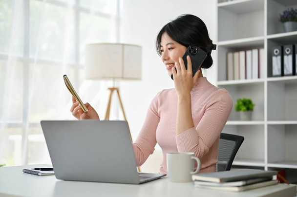 Attraktive asiatische Geschäftsfrau betrachtet die Details auf einem Notizblock, während sie mit jemandem am Schreibtisch telefoniert. - Foto, Bild