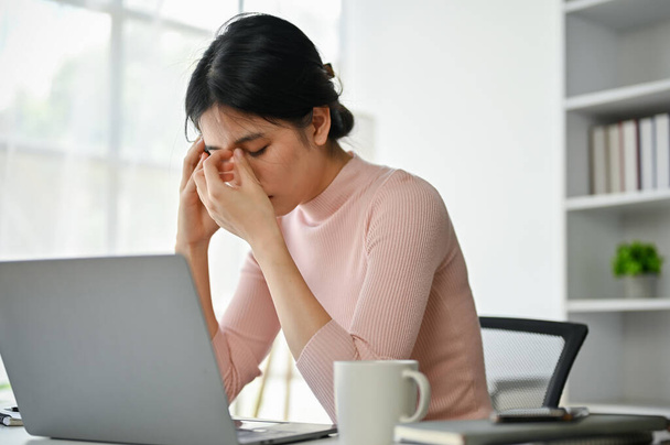 Стрес і втомлена азіатська бізнесменка сидить за своїм столом у своєму кабінеті, страждаючи від головного болю, турбуючись про свою роботу.. - Фото, зображення