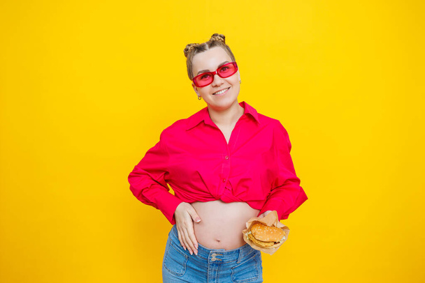 Mujer embarazada alegre en camisa rosa sosteniendo hamburguesa sobre fondo amarillo aislado con sorpresa y expresión facial conmocionada. Alimento nocivo durante el embarazo. Una mujer embarazada come comida rápida - Foto, imagen