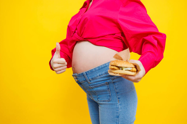 Весела вагітна жінка в рожевій сорочці тримає гамбургер над ізольованим жовтим тлом з несподіваним і шокованим виразом обличчя. Шкідлива їжа під час вагітності. Вагітна жінка їсть фаст-фуд
 - Фото, зображення