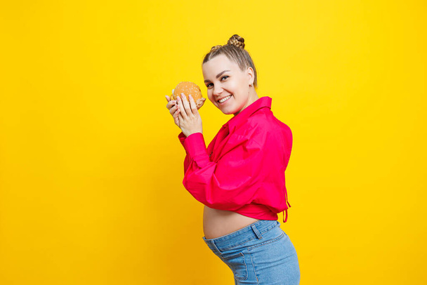 Funny pregnant young woman eating burger. Pregnant woman holding tasty sandwich while standing isolated on yellow studio wall. Junk food concept. Fast food during pregnancy. - Foto, Imagen