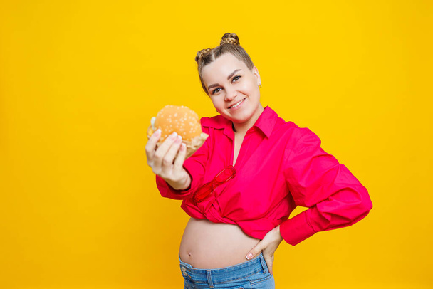 Funny pregnant young woman eating burger. Pregnant woman holding tasty sandwich while standing isolated on yellow studio wall. Junk food concept. Fast food during pregnancy. - Foto, imagen