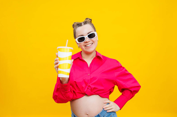 Cheerful pregnant woman in pink shirt holding tasty drink on isolated yellow background. A refreshing drink during pregnancy. A pregnant woman drinks water from a disposable glass - Photo, Image