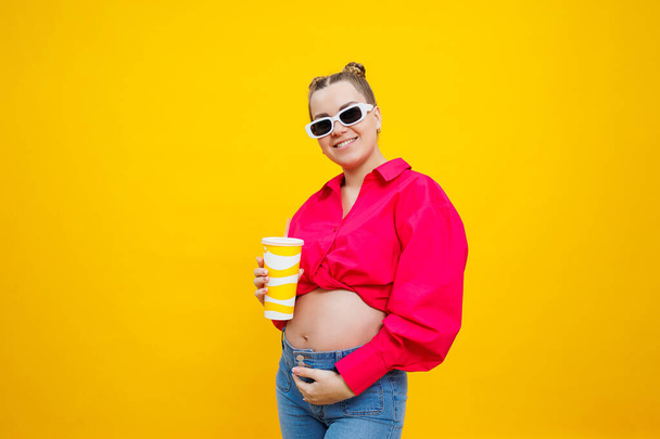 Radosna ciężarna kobieta w różowej koszuli trzymająca smaczny napój na żółtym tle. Odświeżający napój w czasie ciąży. Ciężarna kobieta pije wodę z jednorazowego kieliszka. - Zdjęcie, obraz
