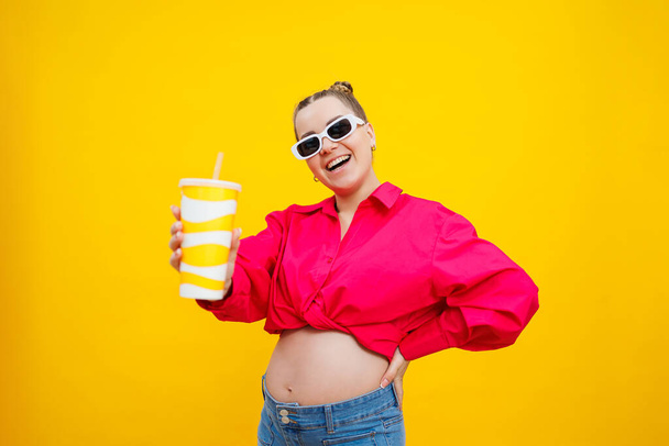 Gut gelaunte Schwangere in rosa Hemd mit leckerem Getränk auf gelbem Hintergrund. Ein erfrischendes Getränk während der Schwangerschaft. Eine Schwangere trinkt Wasser aus einem Einwegglas - Foto, Bild