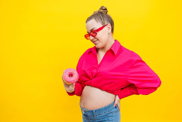 Χαμογελώντας έγκυος γυναίκα σε ροζ πουκάμισο κρατώντας νόστιμα γλυκά ντόνατς σε απομονωμένο κίτρινο φόντο. Γλυκό θερμιδικό φαγητό κατά τη διάρκεια της εγκυμοσύνης. Μια έγκυος γυναίκα τρώει λιπαρά γλυκά. - Φωτογραφία, εικόνα
