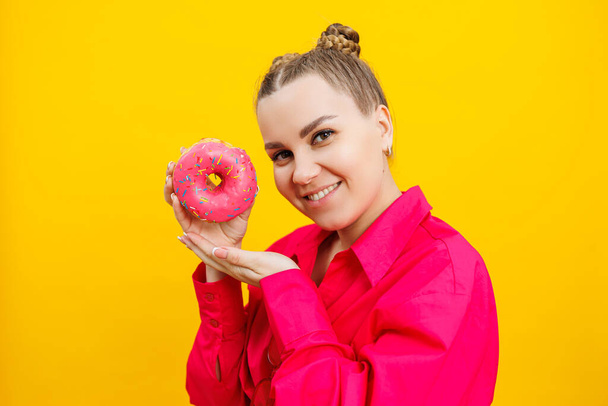 A beautiful pregnant woman holds a sweet donut in her hand on a yellow background. Expecting a child, pregnancy and motherhood. The concept of healthy and unhealthy food, diet. Junk food - Foto, imagen
