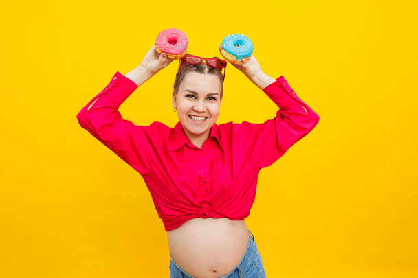 Kaunis raskaana oleva nainen pitää makeaa donitsia kädessään keltaisella pohjalla. Lapsen odottaminen, raskaus ja äitiys. Käsite terveellistä ja epäterveellistä ruokaa, ruokavalio. Roskaruoka - Valokuva, kuva