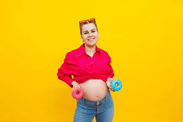 Красива вагітна жінка тримає солодкий пончик в руці на жовтому тлі. Очікування дитини, вагітності та материнства. Концепція здорової та здорової їжі, дієти. Скубічна їжа
 - Фото, зображення