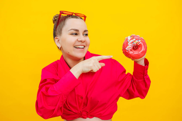 Pregnant young woman in a pink shirt with a donut in her hands. Junk food during pregnancy. Hungry pregnant woman holding donuts while standing on yellow background. - 写真・画像