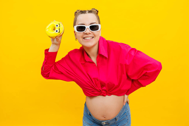 Положительная беременная женщина ест пончики в розовой рубашке на желтом фоне. Счастье от беременности, ожидая ребенка. Высококачественное фото. Вредное питание во время беременности - Фото, изображение