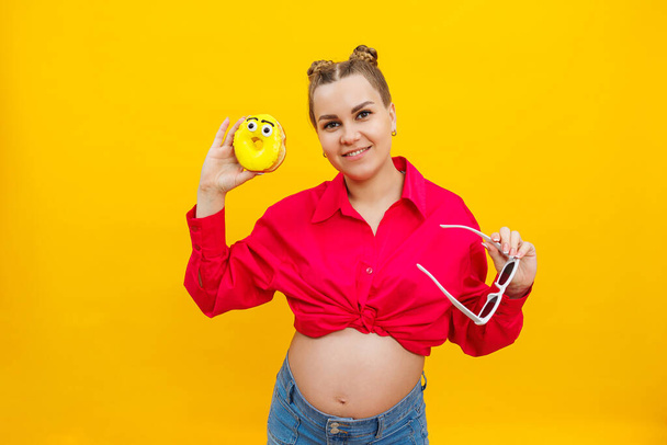 Sarı arka planda elinde tatlı sarı bir çörek tutan neşeli hamile kadın. Bir çocuk, hamilelik ve annelik bekliyor. Sağlıklı ve sağlıksız yiyecek konsepti, diyet. Abur cubur - Fotoğraf, Görsel