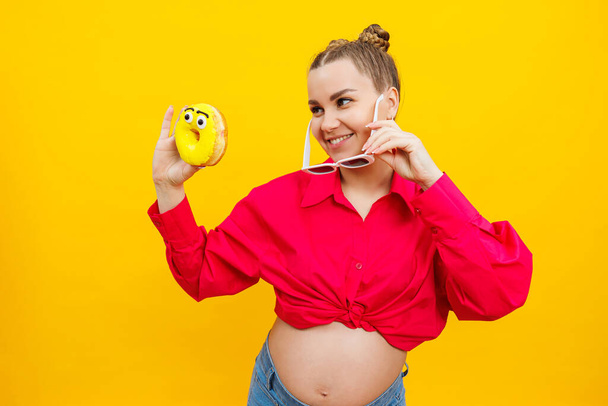 Позитивна вагітна жінка їсть пончики в рожевій сорочці ізольовані на жовтому тлі. Щастя від вагітності, очікуючи дитину. Якісна фотографія. Шкідлива їжа під час вагітності
 - Фото, зображення