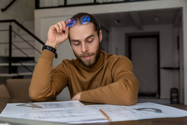 Exhausted tired focused young bearded young engineer architect working on new design of house, creating blueprint, being overworked. - Foto, afbeelding