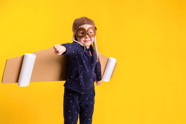 Freedom, girl playing to be airplane pilot, funny little girl with aviator cap and glasses, carries wings made of brown cardboard as an airplane. Studio photography on a yellow background - Foto, immagini