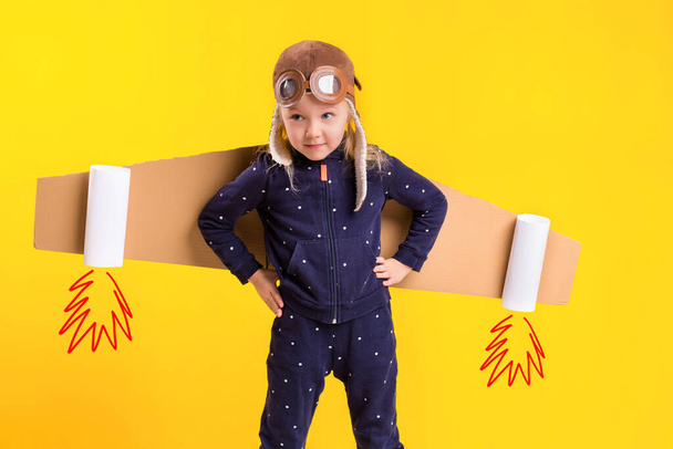 Vrijheid, meisje dat vliegtuigpiloot speelt, grappig klein meisje met vliegeniershoed en bril, draagt vleugels van bruin karton als vliegtuig. Studiofotografie op een gele achtergrond. Verbeelding - Foto, afbeelding