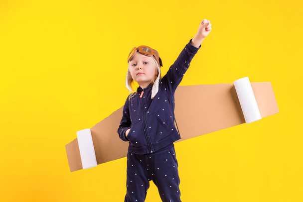 Freedom, girl playing to be airplane pilot, funny little girl with aviator cap and glasses, carries wings made of brown cardboard as an airplane. Studio photography on a yellow background. Imagination - Photo, Image