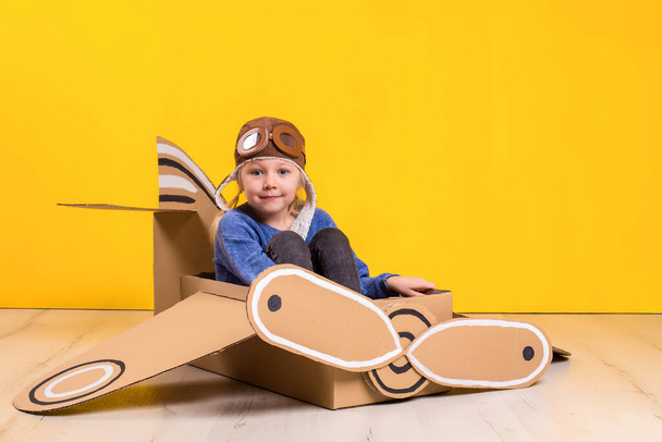 Little dreamer girl playing with a cardboard airplane. Childhood. Fantasy, imagination. Studio photography on a yellow background. Imagination or exploration concept - Фото, зображення