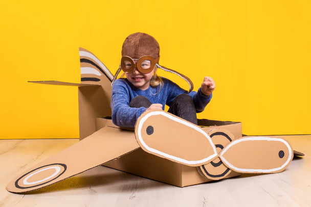 Little dreamer girl playing with a cardboard airplane. Childhood. Fantasy, imagination. Studio photography on a yellow background. Imagination or exploration concept - Zdjęcie, obraz