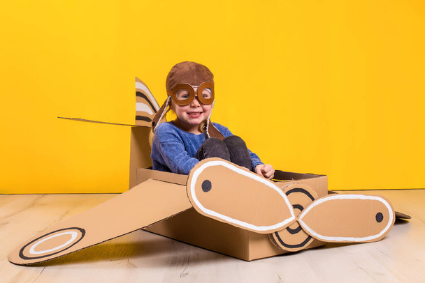 Klein dromer meisje dat met een kartonnen vliegtuig speelt. Jeugd. Fantasie, verbeelding. Studiofotografie op een gele achtergrond. Verbeeldings- of exploratieconcept - Foto, afbeelding