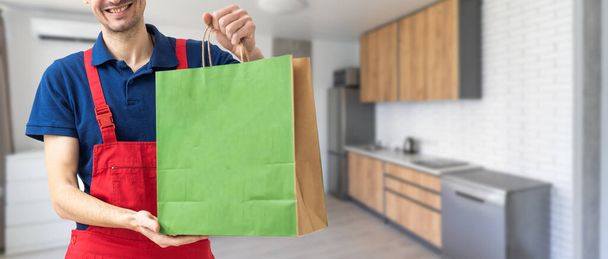Молодой красивый курьер держит бумажный пакет с едой на вынос счастливый с большой улыбкой - Фото, изображение